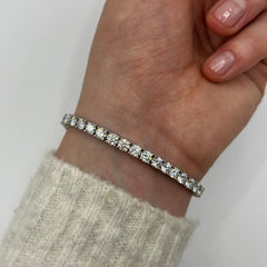 A close-up of a person's wrist wearing a thin silver tennis bracelet with clear round-cut stones, paired with a light beige knit sweater cuff against a plain light background.
