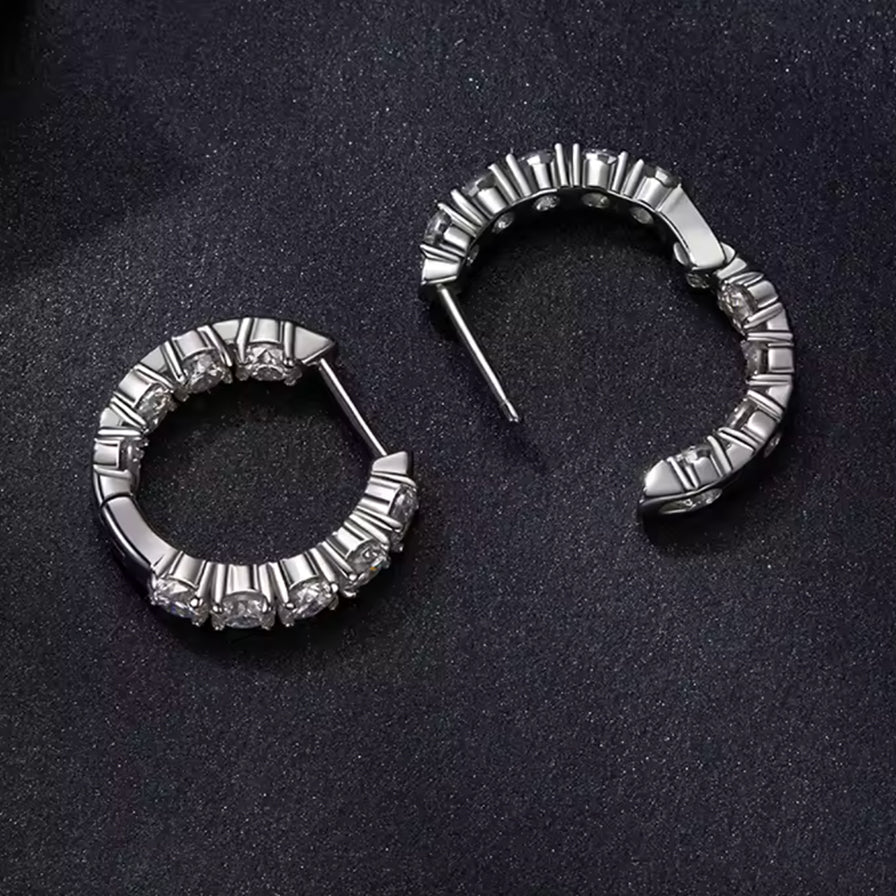 A pair of silver-toned hoop earrings with clear gemstones, displayed on a dark textured background.