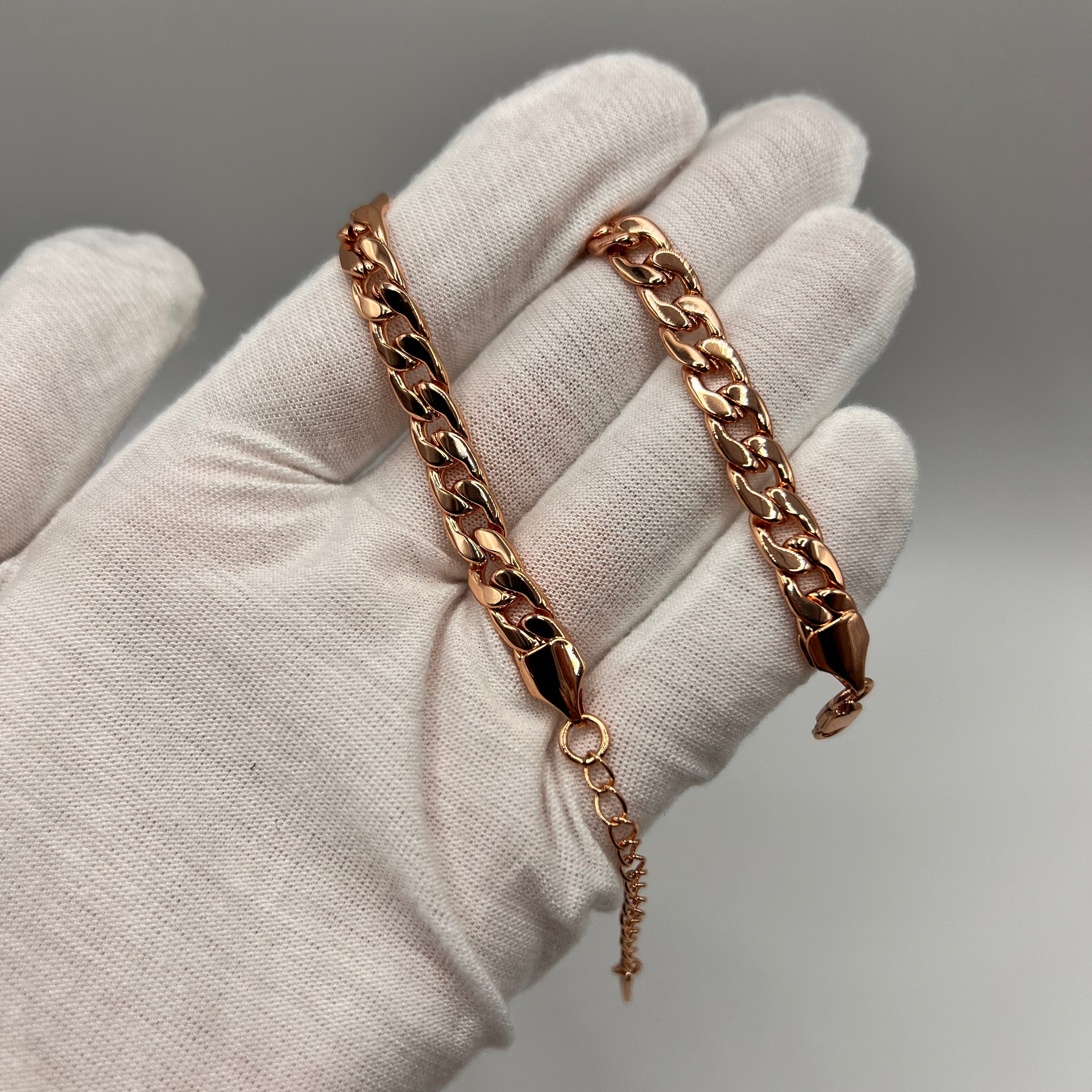 A rose gold curb chain bracelet with a lobster clasp and extender, held in a white fabric-gloved hand against a neutral gray background.
