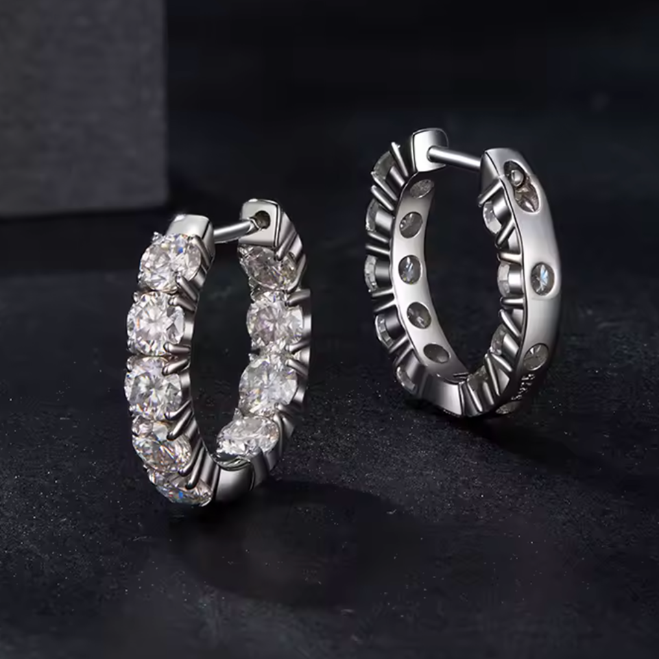 A pair of silver hoop earrings with clear round-cut stones, shown from front and side angles on a dark textured surface.