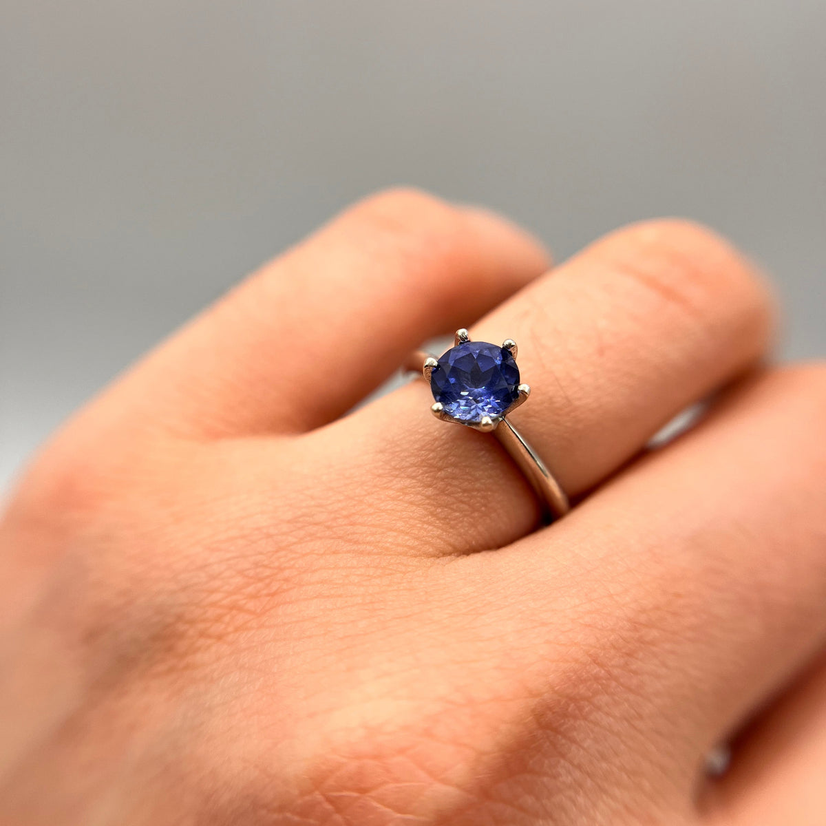 A close-up of a hand wearing a silver ring with a round-cut deep blue gemstone, set against a soft, out-of-focus gray background.