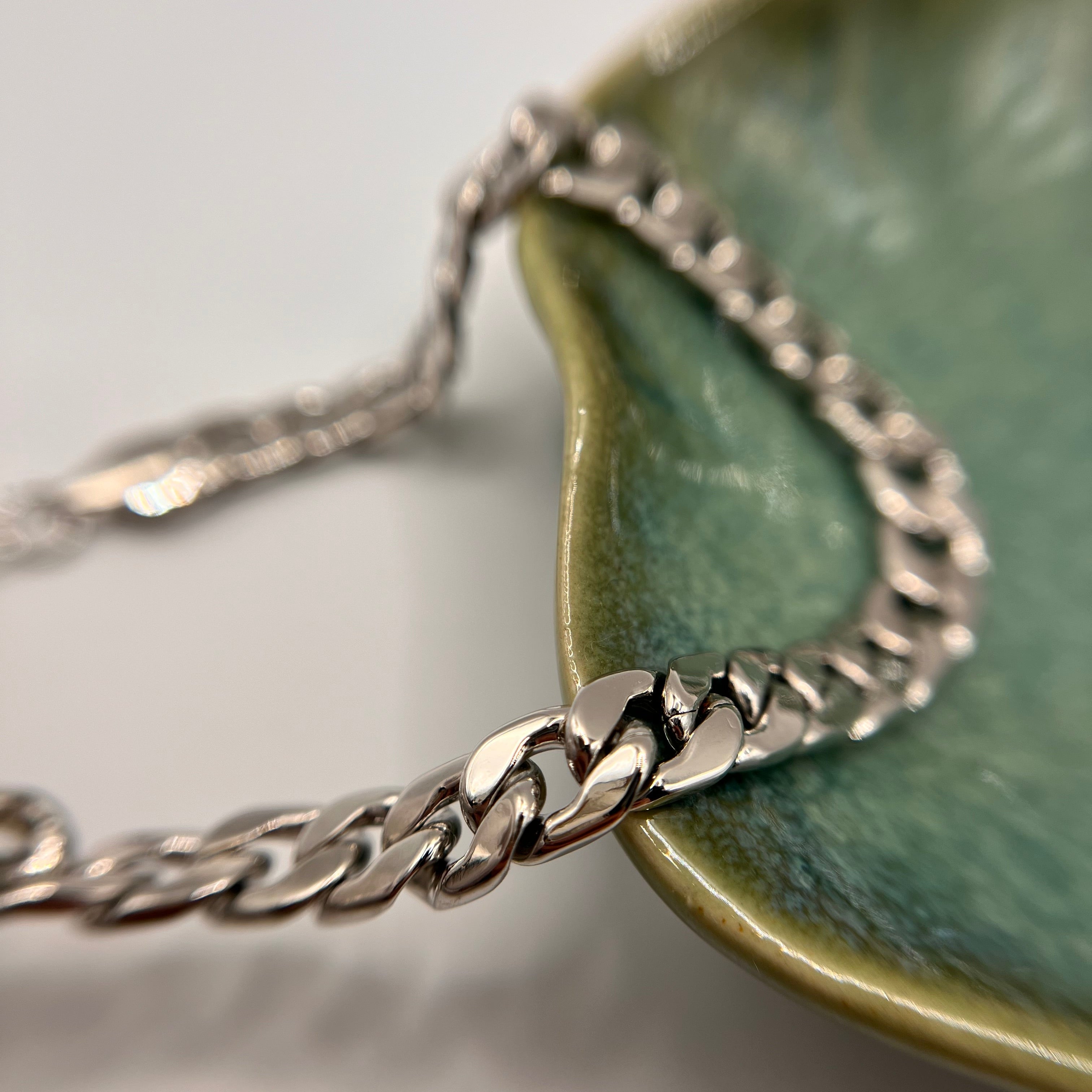 A close-up of a silver-toned curb chain necklace draped over a glossy green ceramic dish on a light gray background.