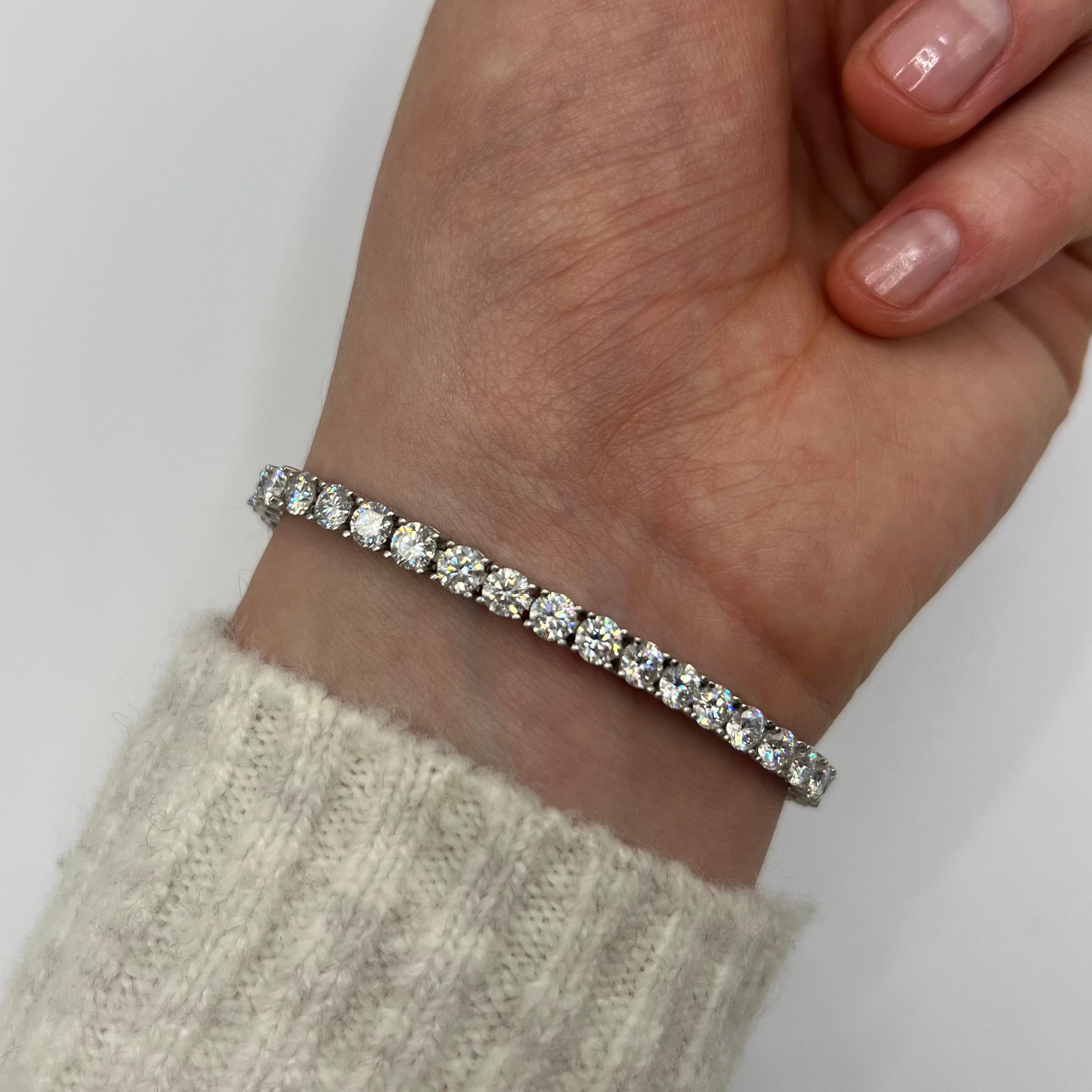 A close-up of a person's wrist wearing a thin silver tennis bracelet with clear round-cut stones, paired with a light beige knit sweater cuff against a plain light background.