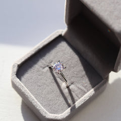 A fine moissanite ring featuring a shiny pure silver band, resting in a plush grey velvet gift box, with the stone's rainbow brilliance on full display