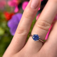A close-up of a hand wearing a solitaire ring with a round-cut deep blue gemstone in a silver-toned setting, with a blurred background of colorful flowers.