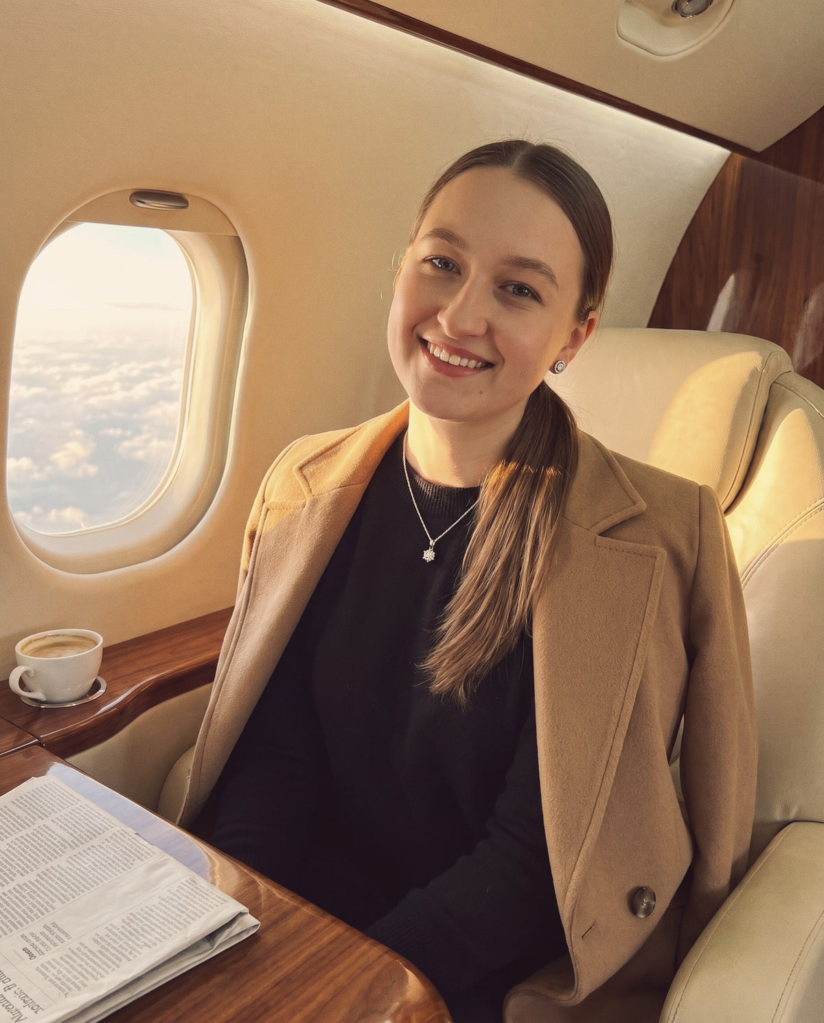 Woman smiling in a private jet cabin in a camel coat, wearing a moissanite pendant necklace