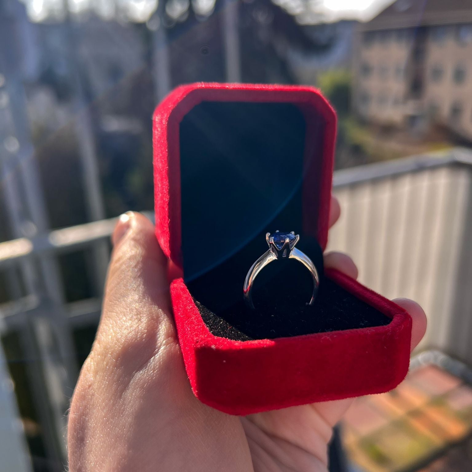 Red velvet box with a silver ring inside, held by a hand on a balcony.